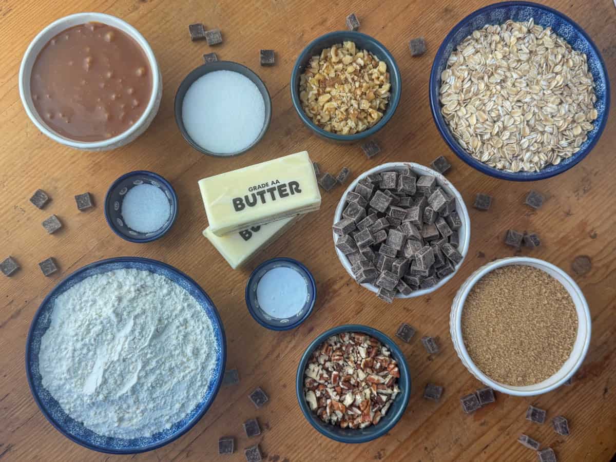 All ingredients needed for Carmelitas bars: flour, white sugar, brown sugar, oats, pecans, walnuts, caramel sauce, chocolate chips or chunks, butter, baking soda and salt