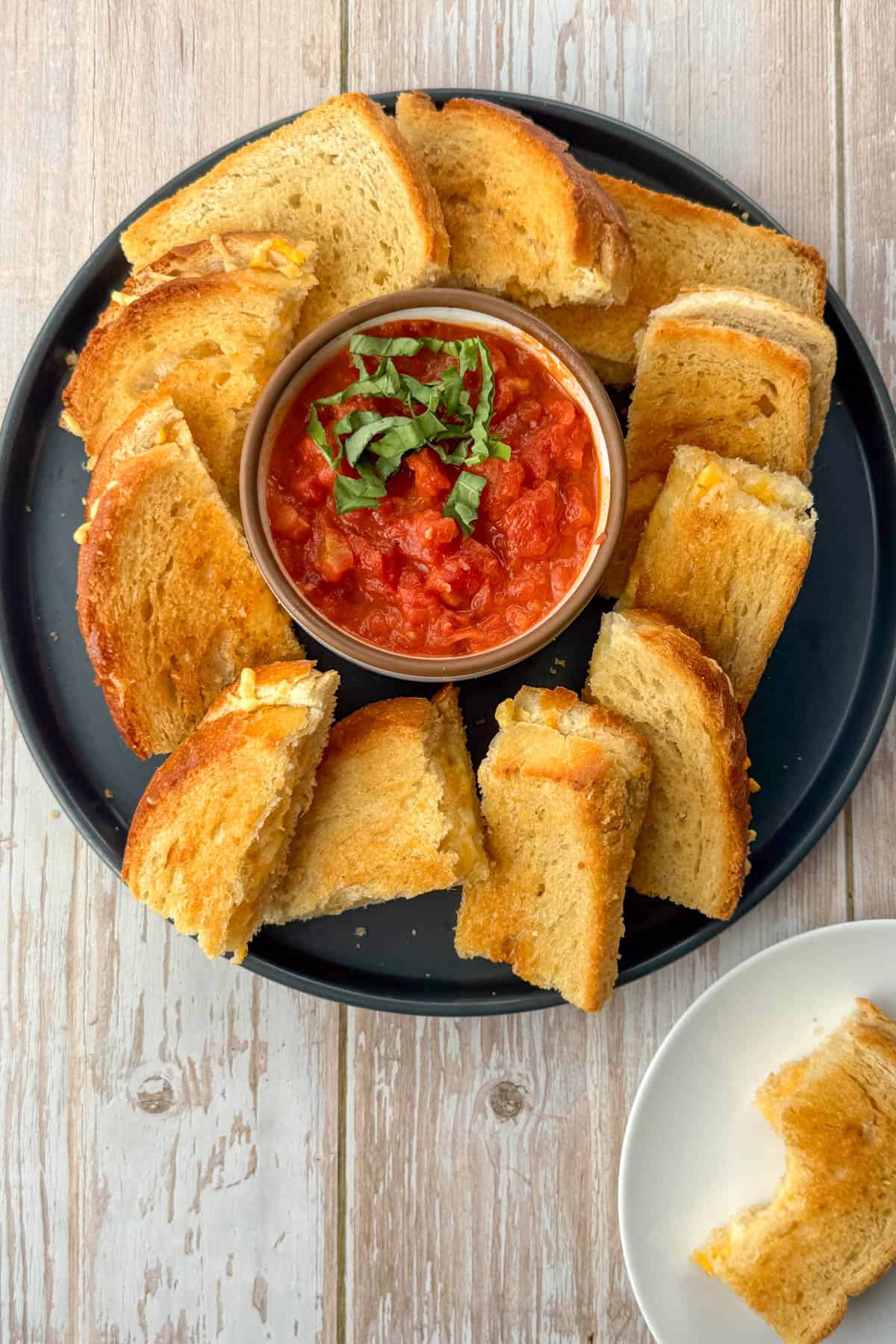 Grey plate with appetizer size pieces of baked grilled cheese sandwiches with tomato dipping sauce with white appetizer plate with grilled cheese sandwich with bite taken out