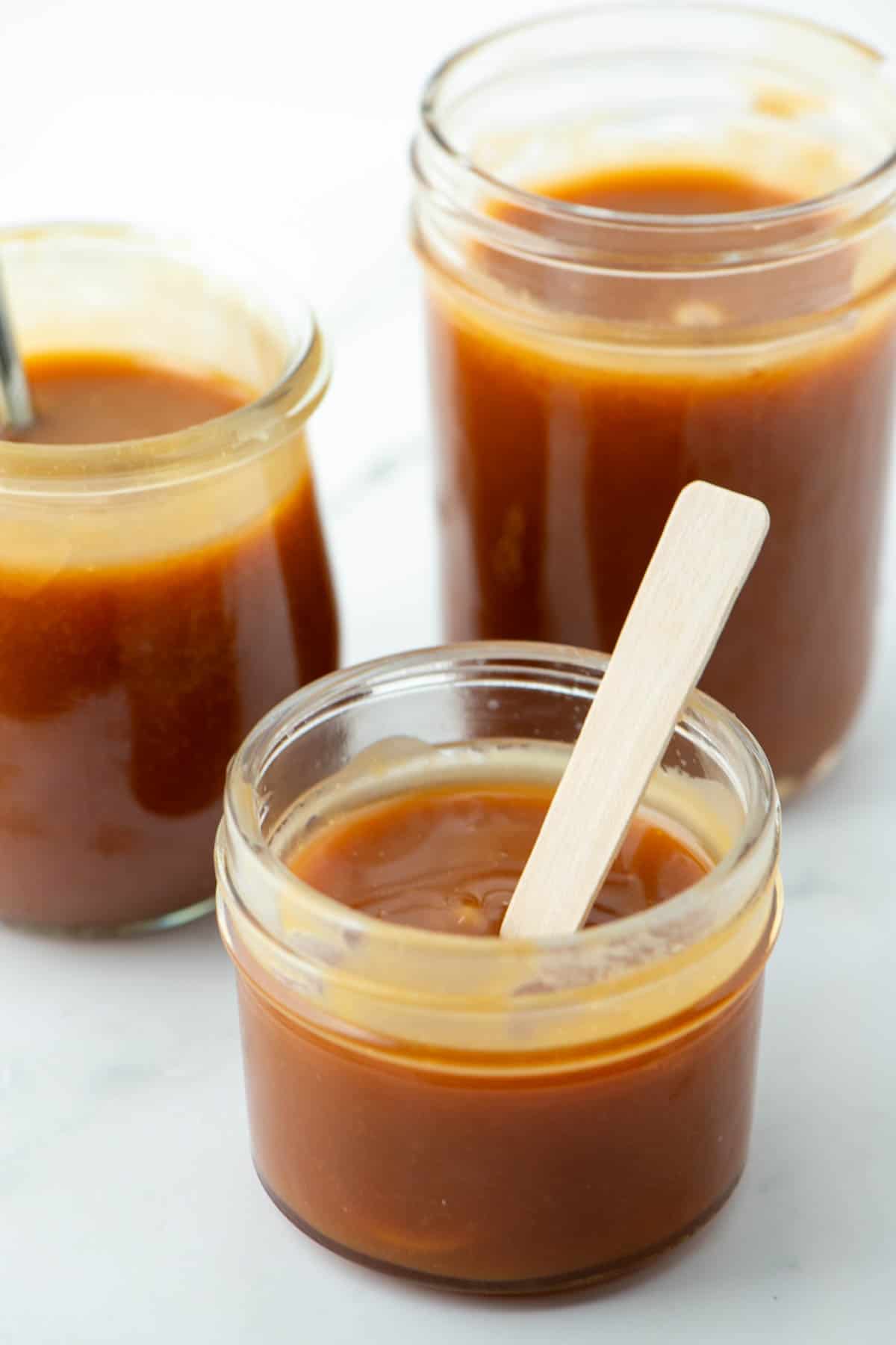 Three different sized small jars of homemade salted caramel sauce. The jar in front has a wooden spoon in it.