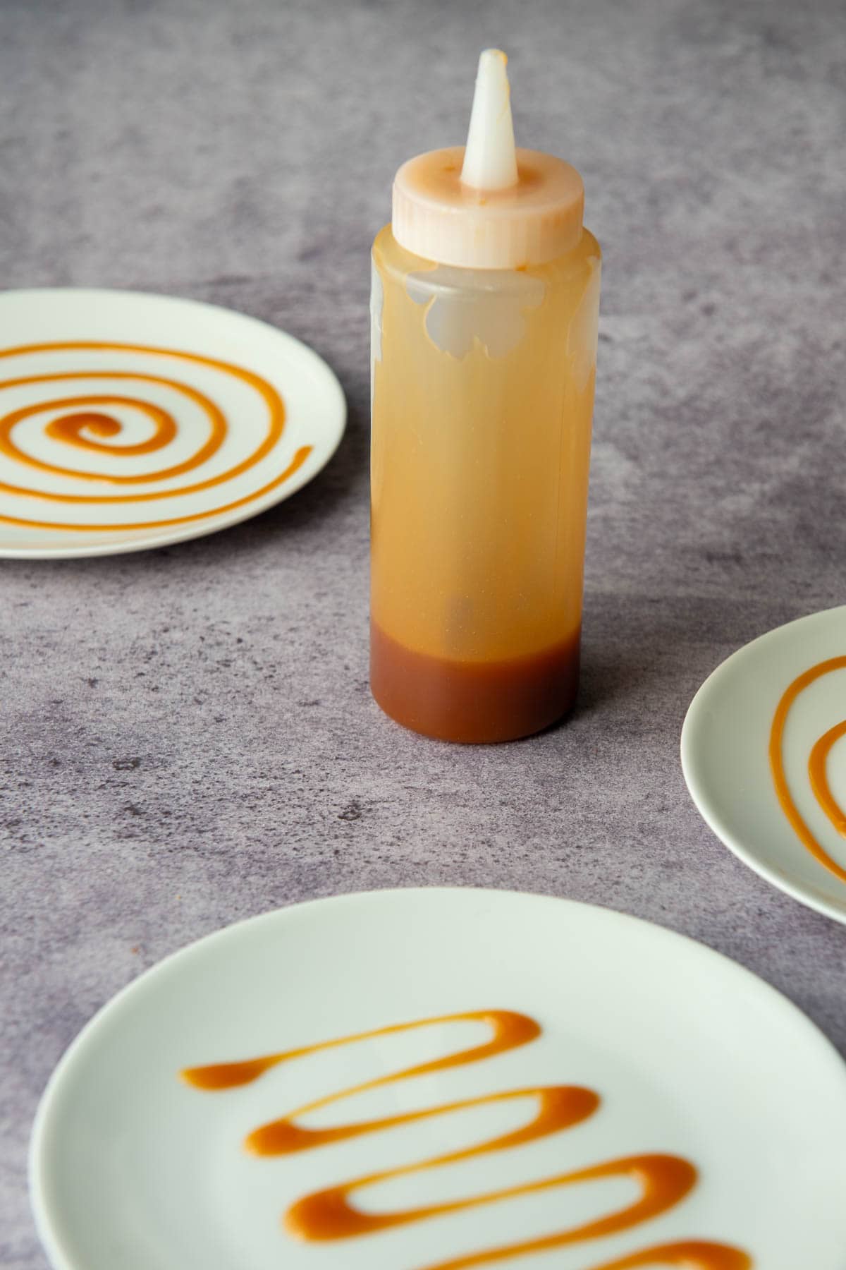 Decorator bottle filled with salted caramel sauce with three plates in partial view that have swirly designs made with the salted caramel on them