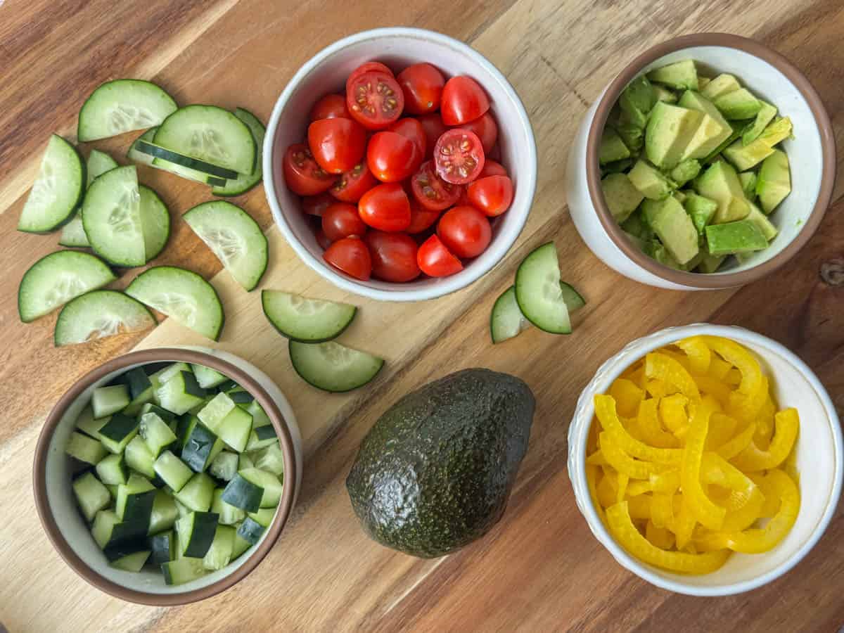 Photo of fruit choices for making a salad: sliced cucumbers, diced cucumbers, halved cherry tomatoes, diced avocado, and sliced yellow bell peppers