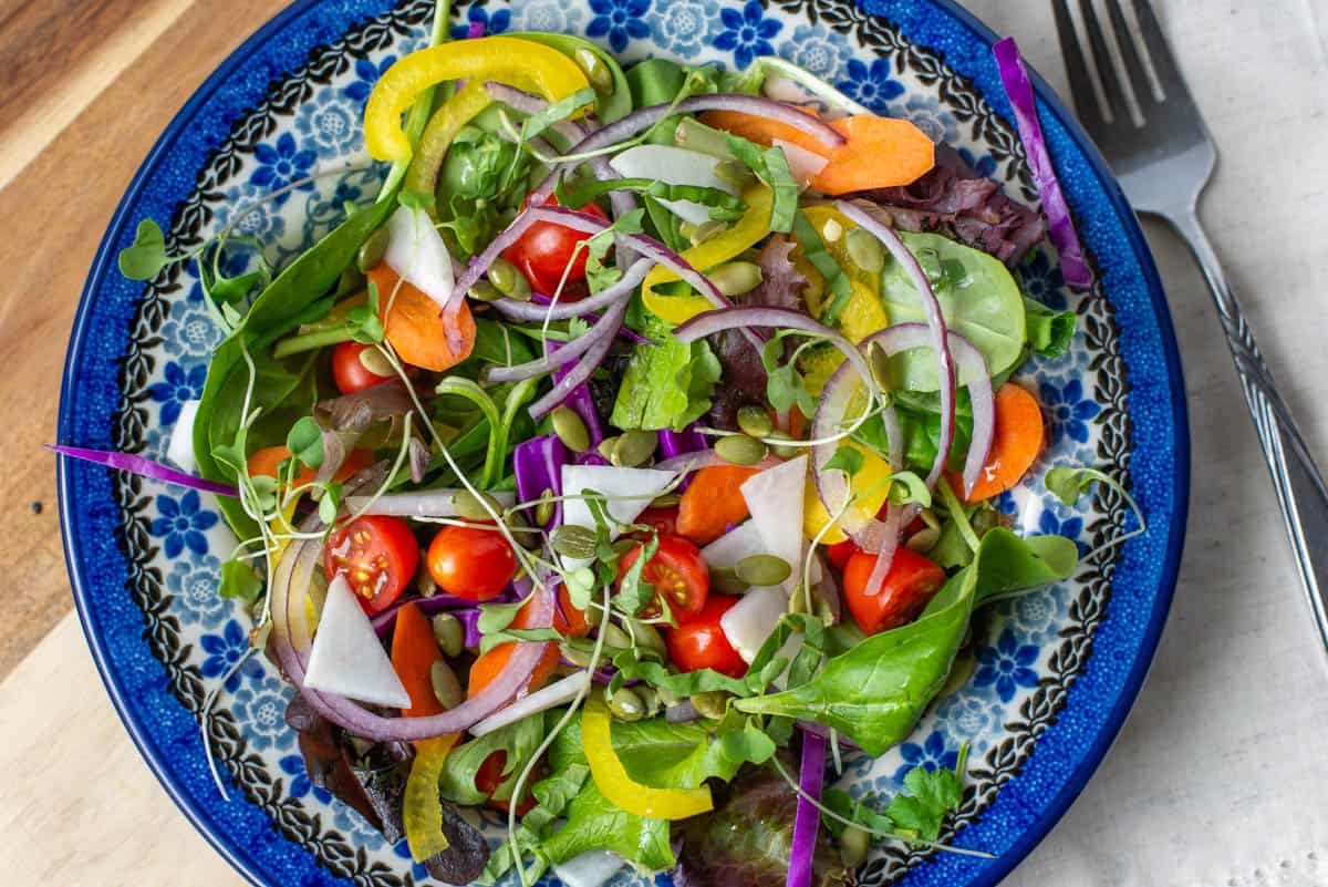 Photo of salad on blue salad bowl. Salad is topped with tomatoes, red cabbage, turnips, carrots, micro greens, basil, pipits seeds, oil and lemon juice.