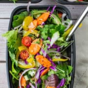 Photo of salad in meal prep container with fork with two meal prep containers with salad in the background. Salad is topped with purple cabbage, cucumbers, carrots, tomato, yellow bell pepper, micro greens, sunflower seeds, sesame seeds and dill.