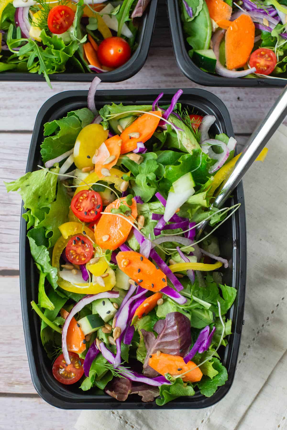 Photo of salad in meal prep container with fork with two meal prep containers with salad in the background. Salad is topped with purple cabbage, cucumbers, carrots, tomato, yellow bell pepper, micro greens, sunflower seeds, sesame seeds and dill.