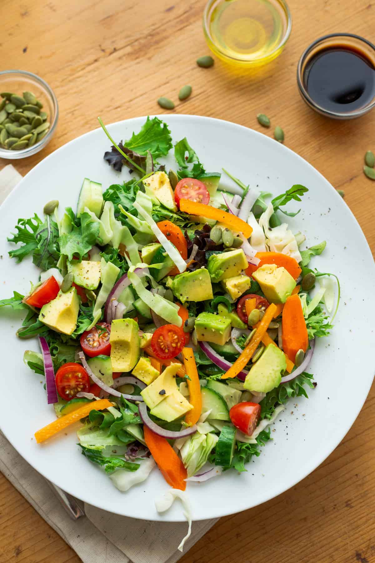 Photo of salad on plate. Salad is topped with green cabbage, red onions, tomatoes, carrots, orange bell pepper, avocados, pepita seeds, cilantro, pepper, soy sauce and olive oil. Little bowls of pepita seeds, oil and soy sauce in the background.