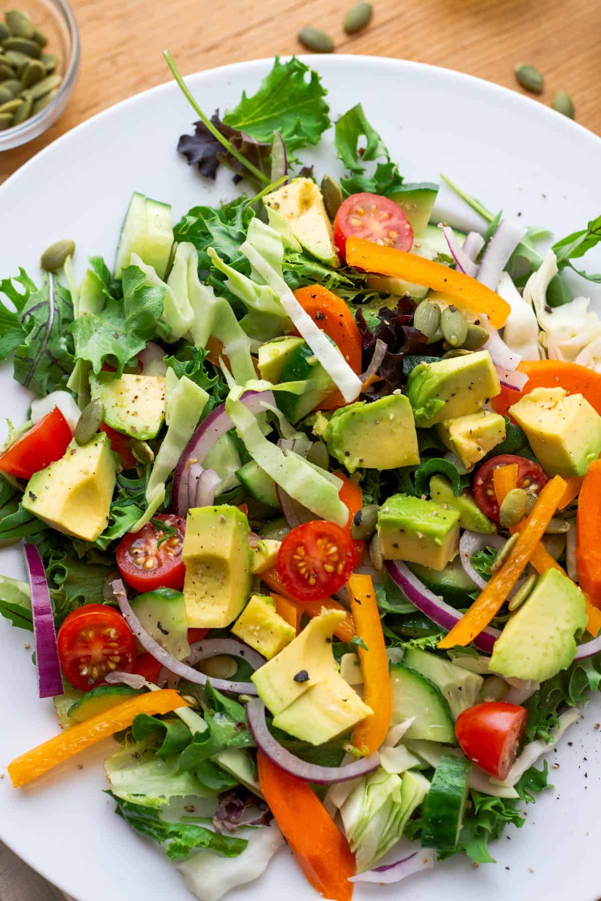 Close up photo of salad on plate. Salad is topped with green cabbage, red onions, tomatoes, carrots, orange bell pepper, avocados, pepita seeds, cilantro, pepper, soy sauce and olive oil