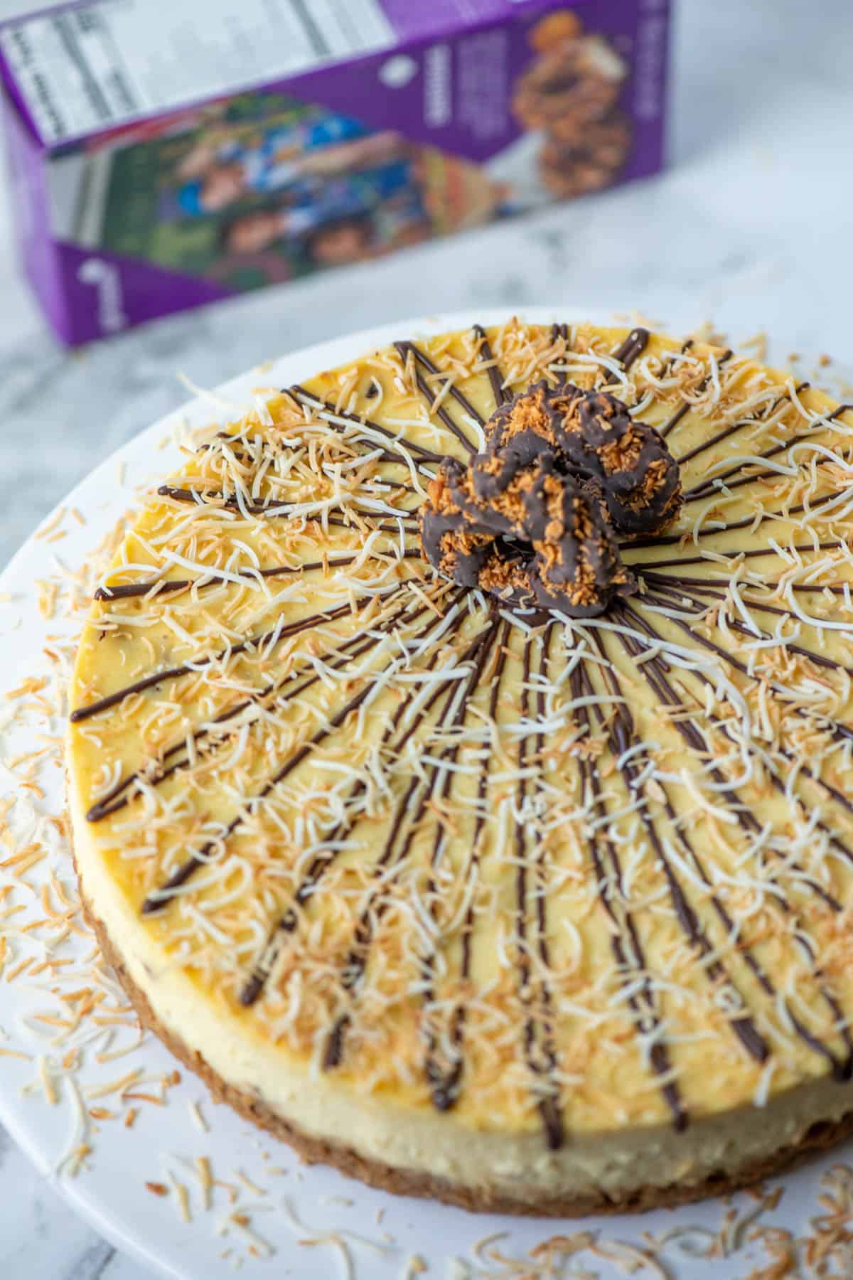 Overhead view of full Samoas cheesecake topped with chocolate, toasted coconut and Samoas cookies with box of Samoas cookies blurred in the background