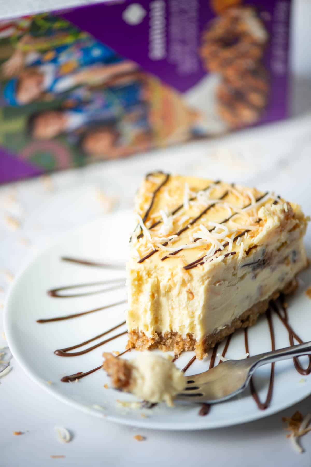 Slice of Samoas cheesecake topped with chocolate and toasted coconut on a plate with a bite of cheesecake on a fork on the plate with a box of Samoas cookies in the background.