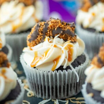 Close up of Samoas cupcakes on a plate with toasted coconut and a box of Samoas cookies blurred in the background