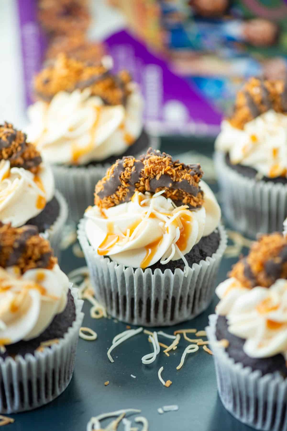 Six Samoas cupcake on a plate with toasted coconut and a box of Samoas cookies blurred in the background