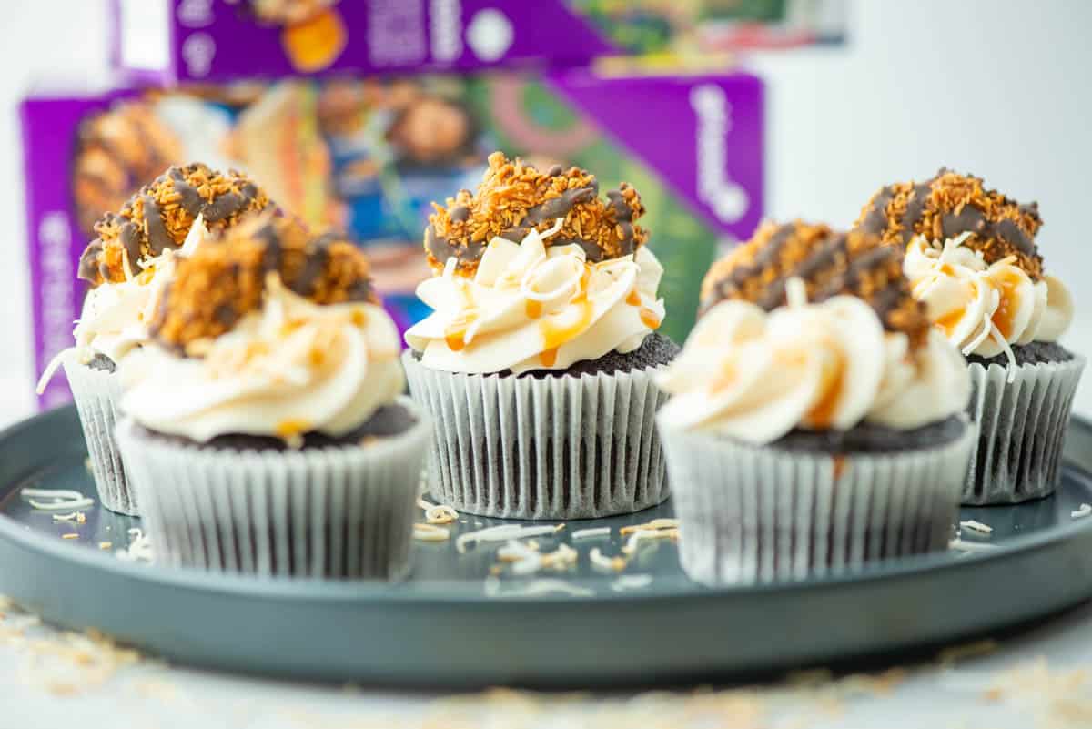 5 Samoas cupcakes on a plate with toasted coconut and boxes of Samoas cookies blurred in the background