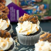 Four Samoas cupcakes on a plate with a box of Samoas cookies blurred in the background