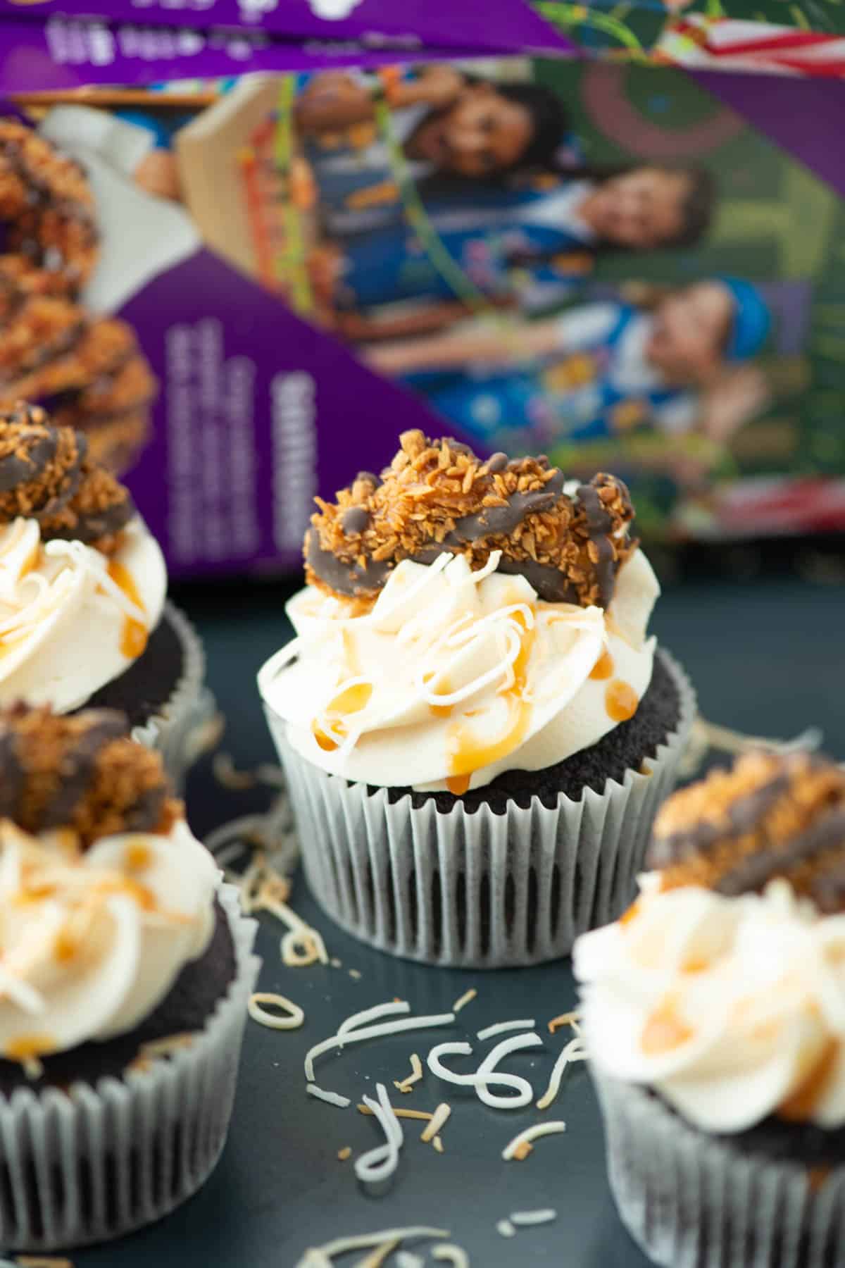 Four Samoas cupcakes on a plate with a box of Samoas cookies blurred in the background