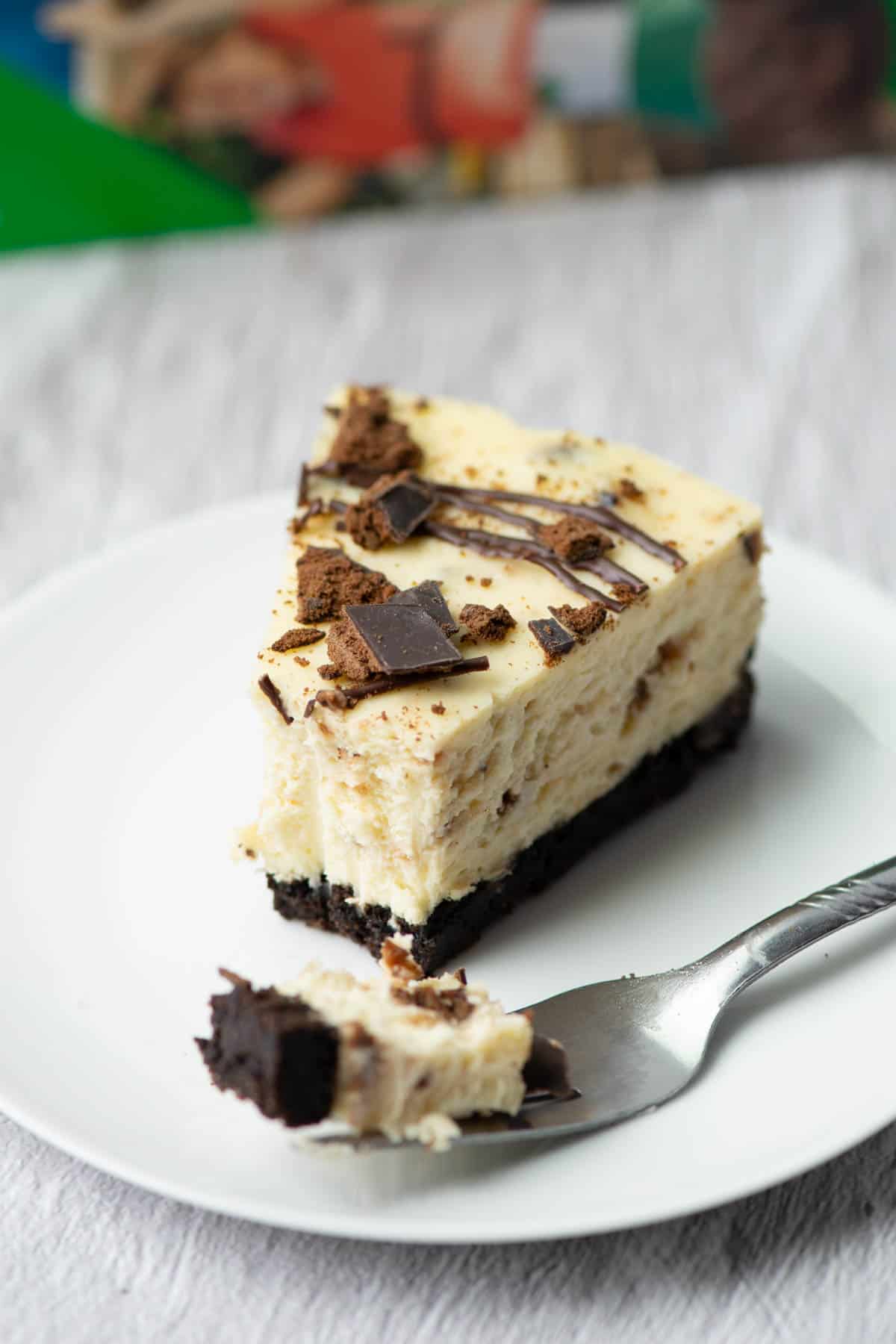 One slice of Thin Mints cheesecake on a plate with a bite on a fork in front with a box of Thin Mints in the background