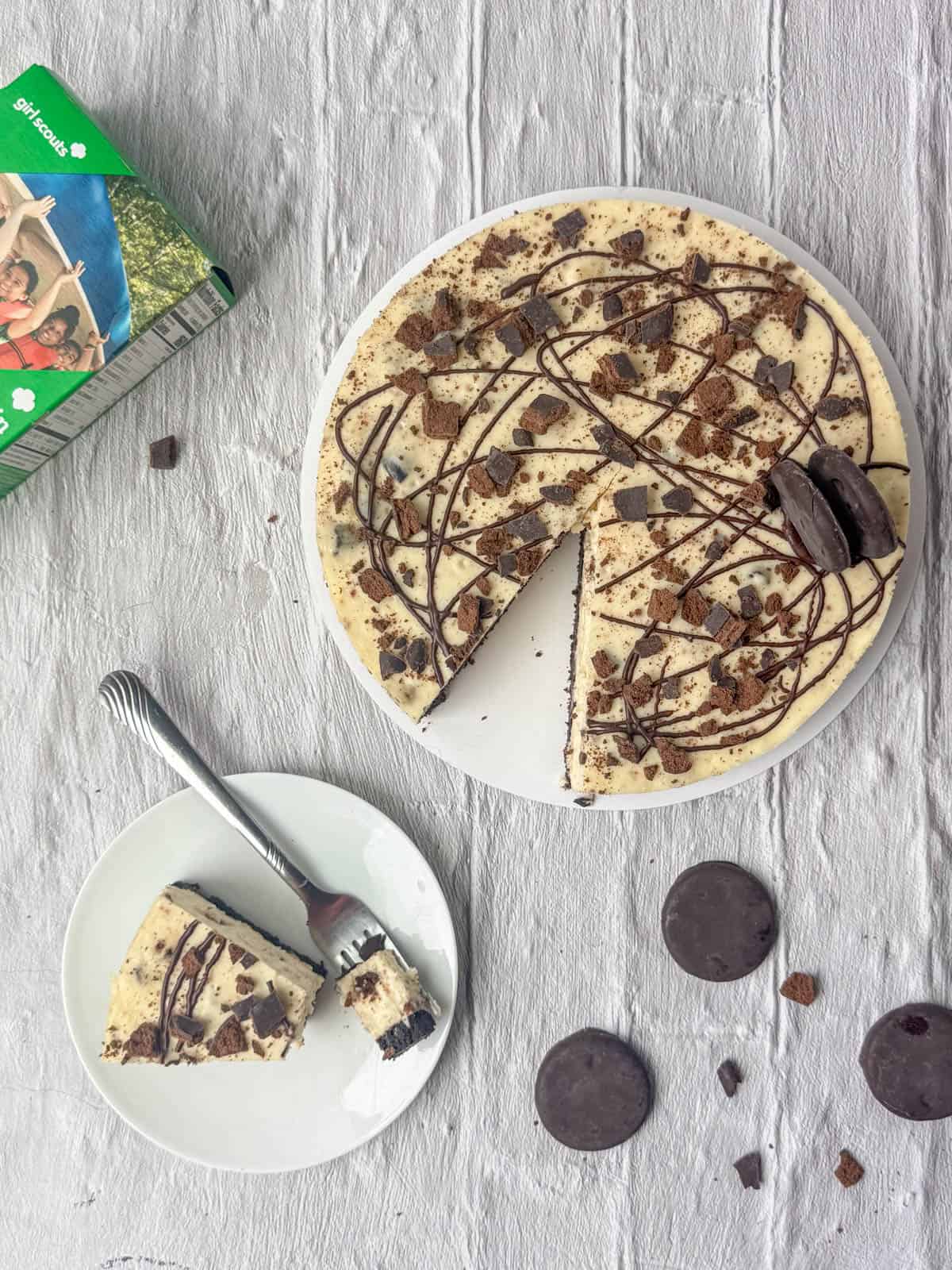 Overhead view of Thin Mints cheesecake on a cakeboard plate with a slice of Thin Mints cheesecake on another plate with three Thin Mints cookies and a box of Thin Mints nearby