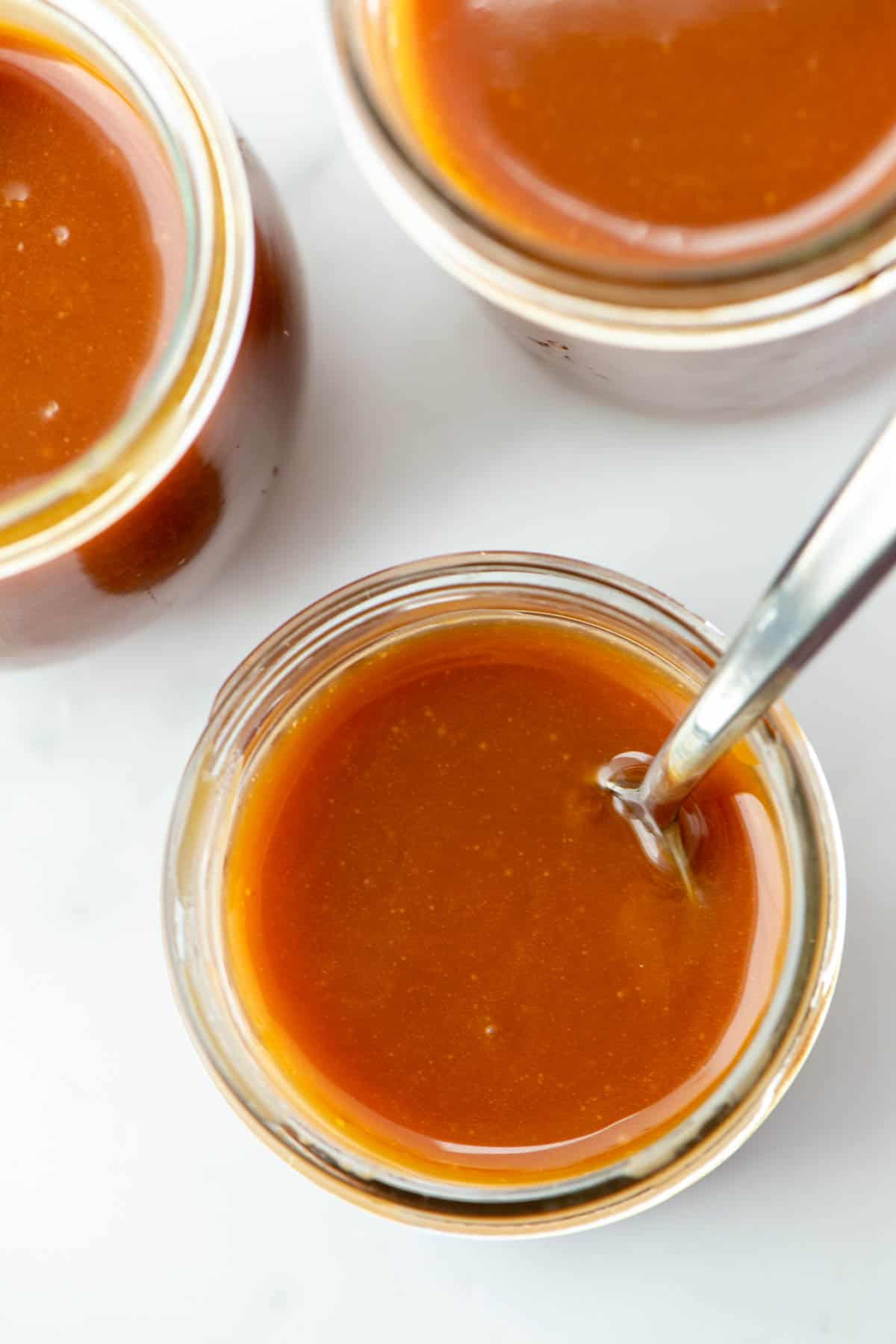 Overhead view of three different sized small jars of homemade salted caramel sauce. The jar in front has a spoon in the jar.