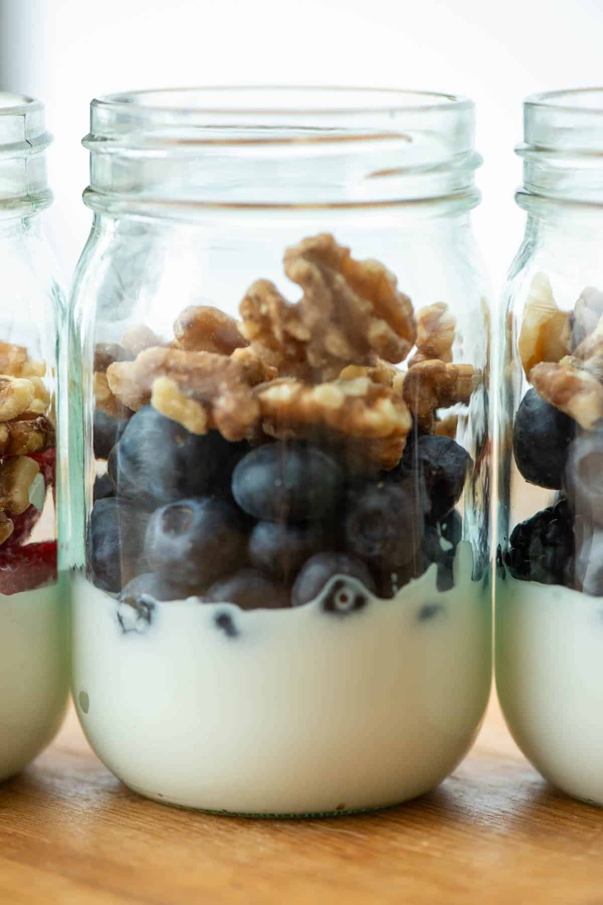 Side view of mason jar filled with greek yogurt, blueberries and walnuts next to two other jars filled with berry yogurt parfait partially visible