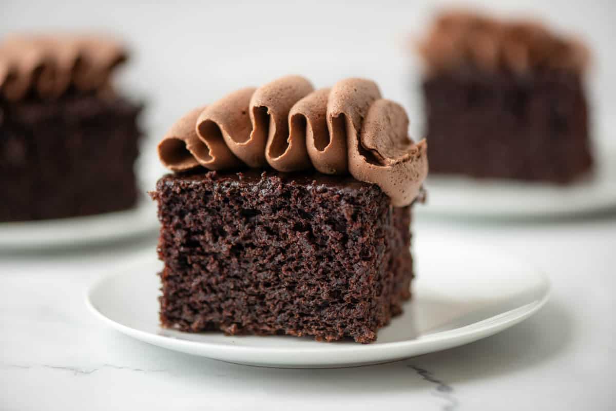 Close up photo of a slice of chocolate zucchini cake with chocolate buttercream frosting. Two additional slices of cake with frosting in the background.