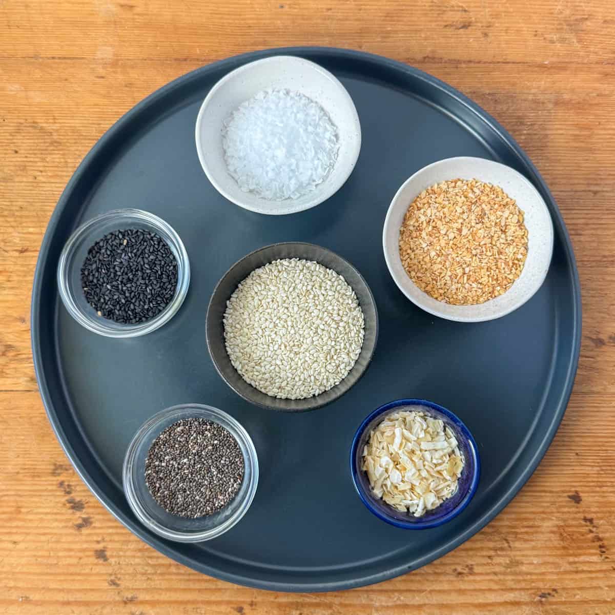 Photo of ingredients needed to make everything but bagel seasoning in tiny bowls: white sesame seeds, flakes salt, dried granulated minced garlic, dried minced onion, black sesame seeds and poppy seeds