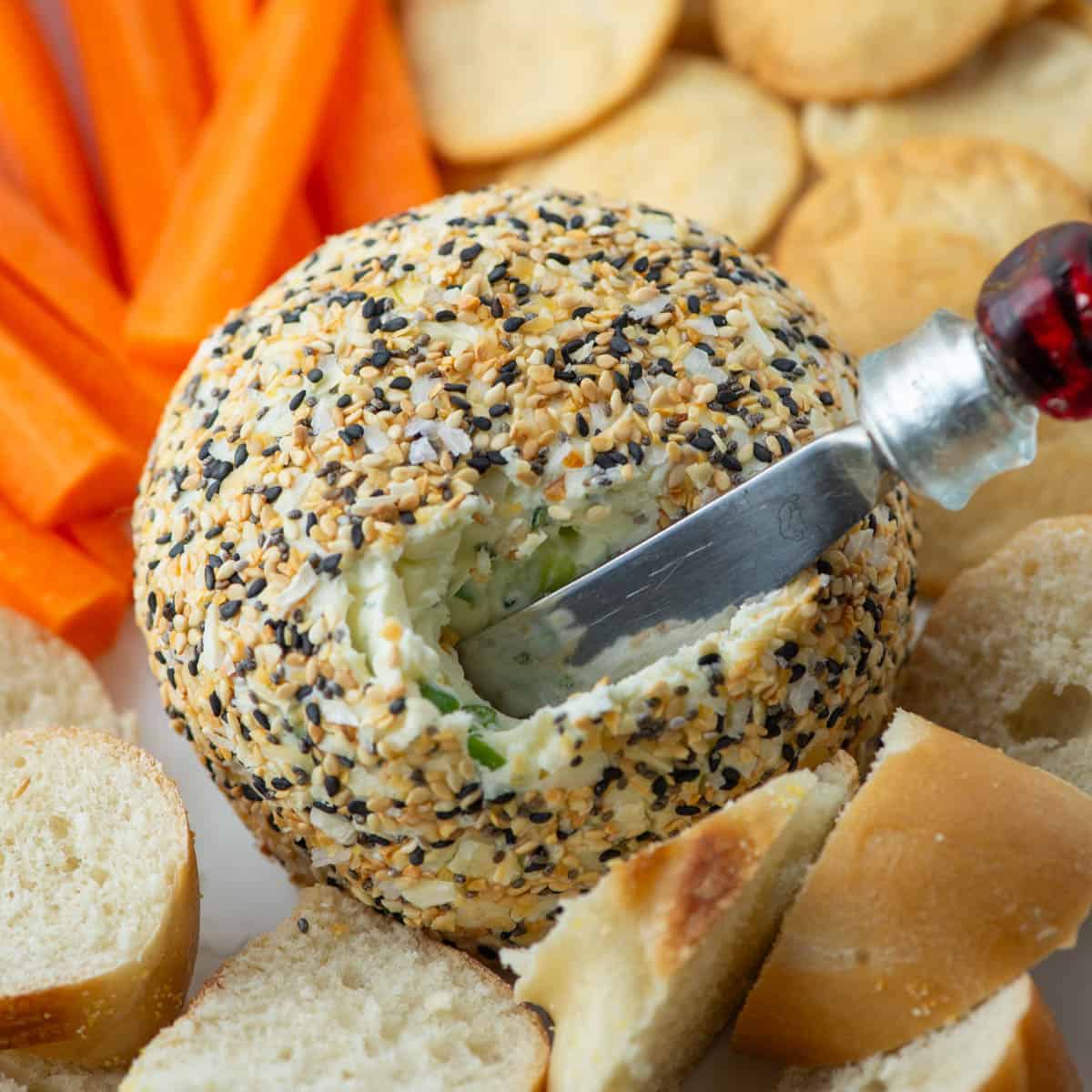 Close up photo of Everything Bagel Cheeseball with a cheese knife spreader sticking out of it surround by plain bagel slices, carrots and pita crackers