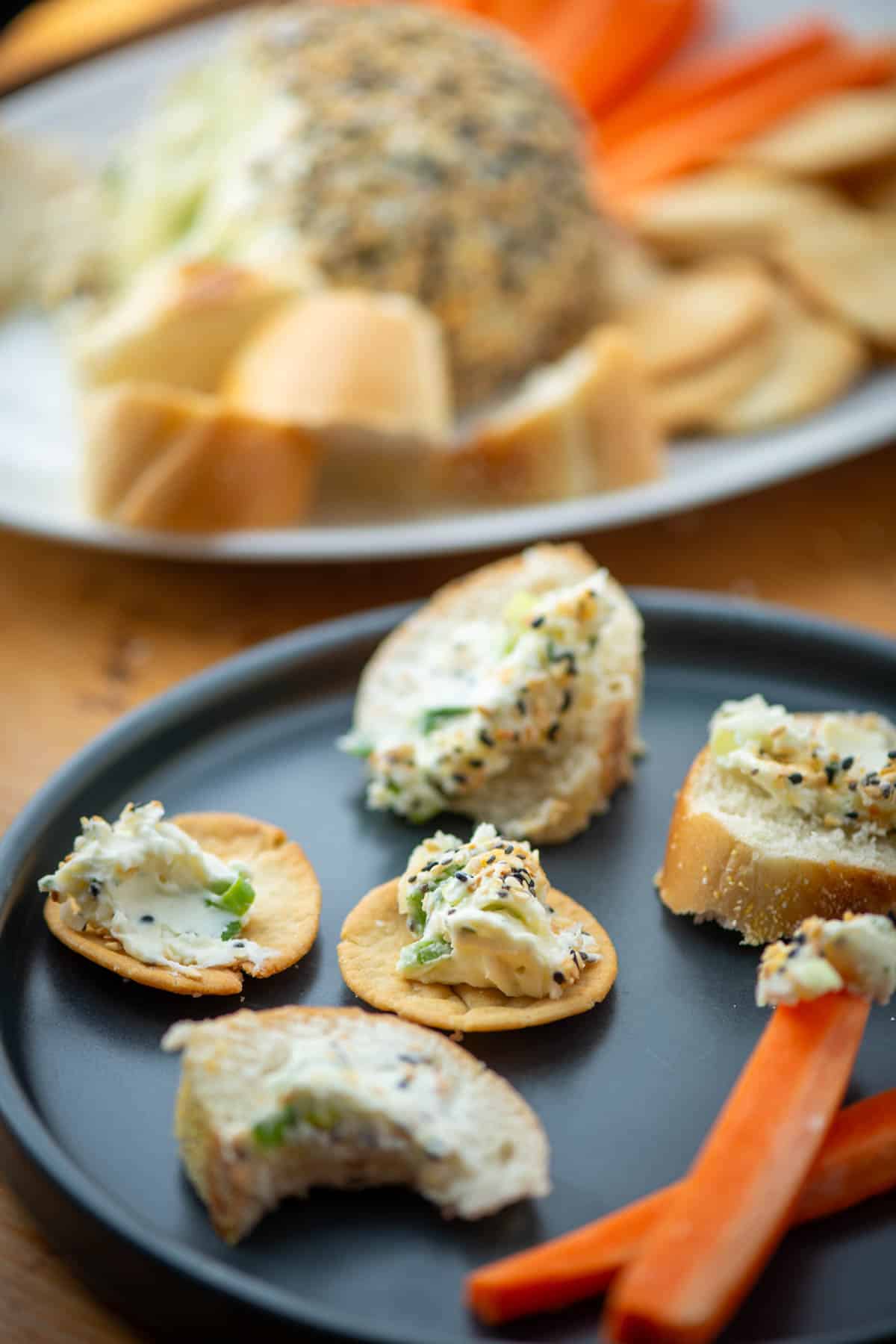 Plate with small bites of sliced bagels, pita chips and carrots with everything but bagel seasoning cheeseball on each bite in front of the serving platter with the everything but bagel seasoning cheeseball and extra bagel slices, pita chips and carrots