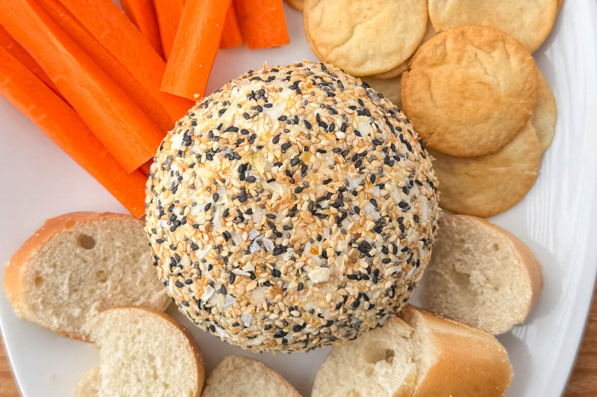 Close up photo of an everything bagel cheeseball on a serving platter surrounded by plain bagel slices, carrots and pita chips