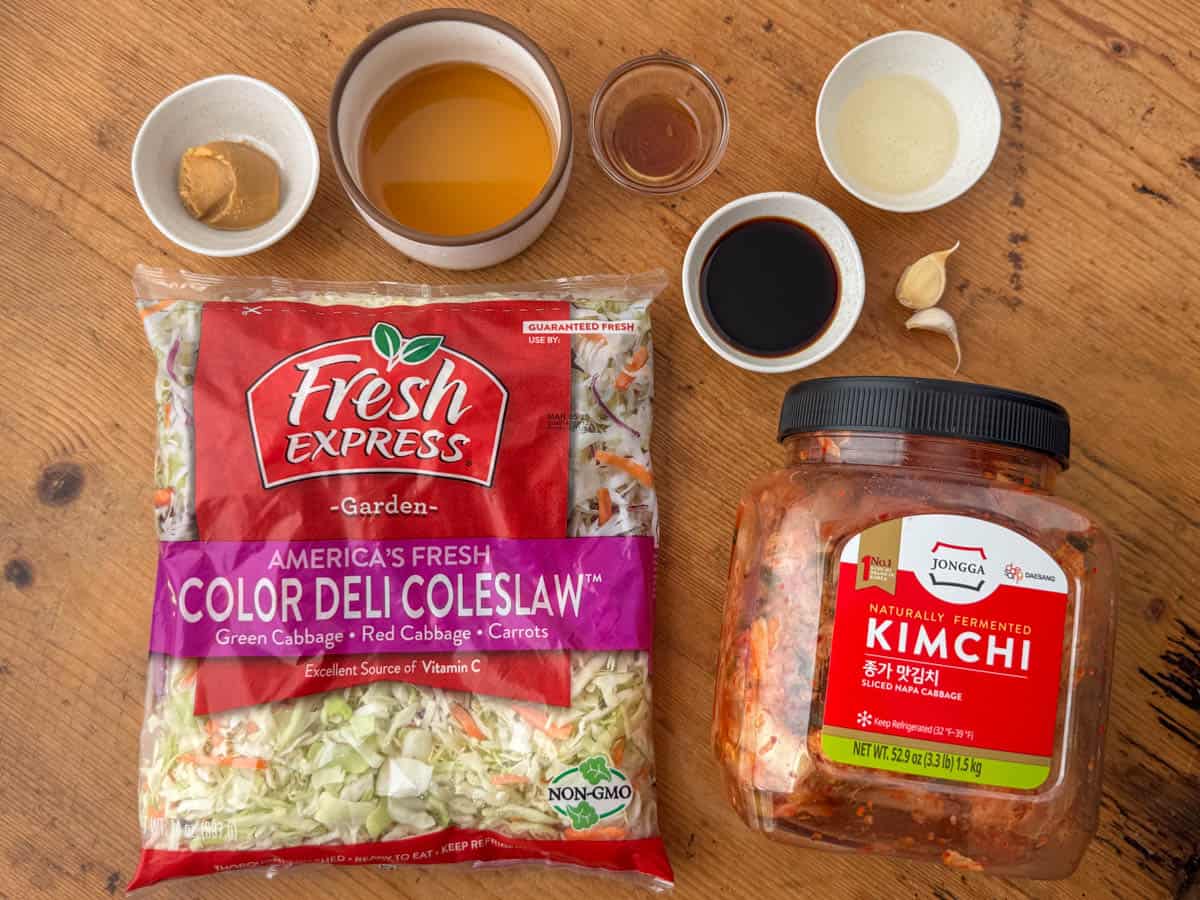 Ingredients needed to make kimchi slaw: prepackaged coleslaw cabbage, kimchi, sesame oil, soy sauce, rice vinegar, white miso, garlic and maple syrup