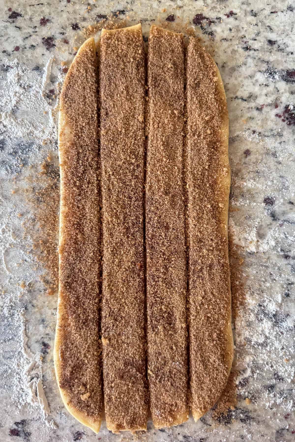 Cinnamon roll dough rolled out into rectangle topped with softened butter, brown sugar and cinnamon and cut into 4 strips ready to be rolled.