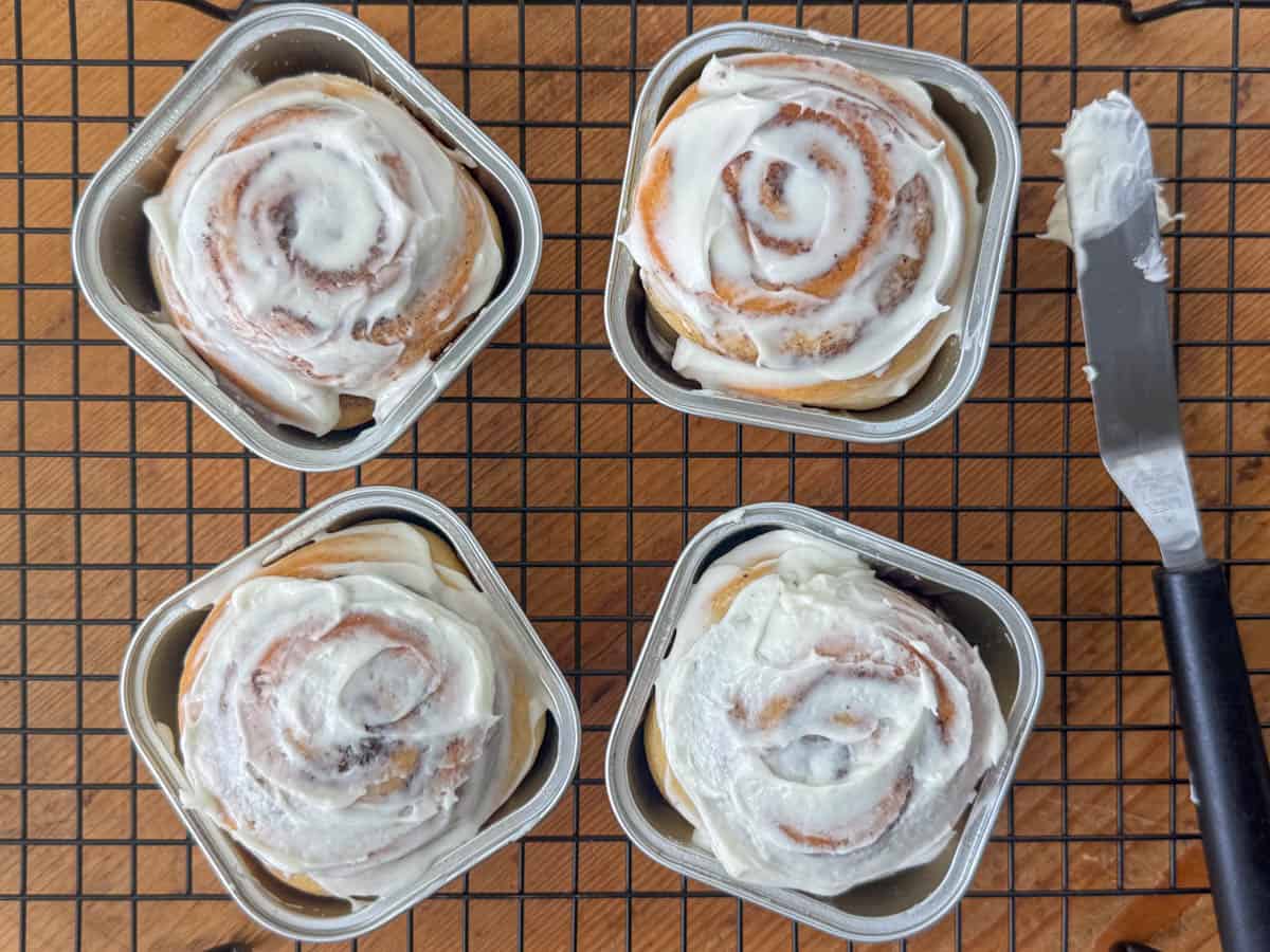 Overhead view of four cinnamon rolls in mini square baking cups next to an icing spatula with cream cheese frosting on it on top of a cooling rack.