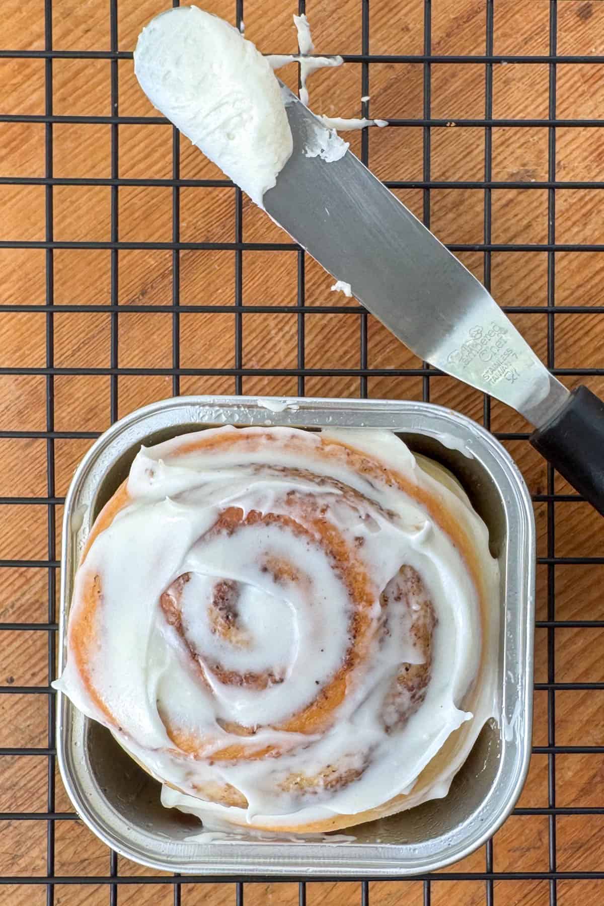 Overhead close up photo of a cinnamon roll in a mini square baking cup next to an icing spatula with cream cheese frosting on it.