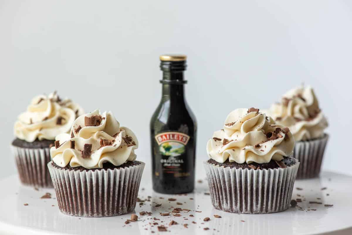 Four chocolate Baileys Irish Cream cupcakes with Baileys buttercream frosting on a cake plate with a mini bottle of Baileys Irish Cream in the midst of the cupcakes