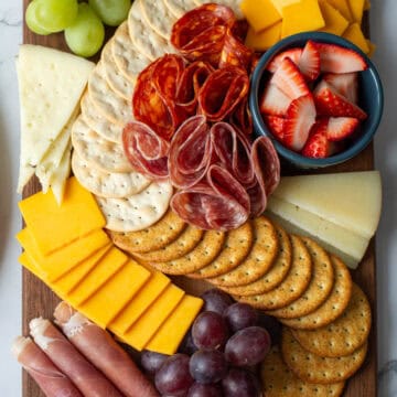 Top view of charcuterie board with cheddar cheese, manchego cheese, assorted crackers, salami, prosciutto, strawberries, green grapesand red grapes