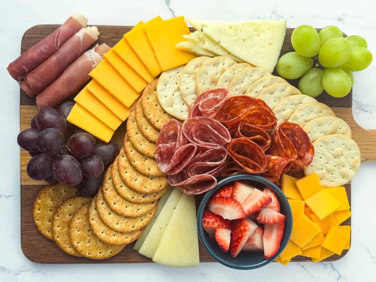 Top view of finished charcuterie board with assorted crackers, assorted meats, assorted cheeses, red and green grapes, and cut strawberries