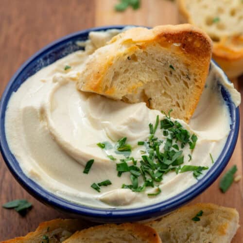 Close up photo of a bowl of whipped tofu with shopped parsley garnish and a piece of garlic toast dipping into the bowl