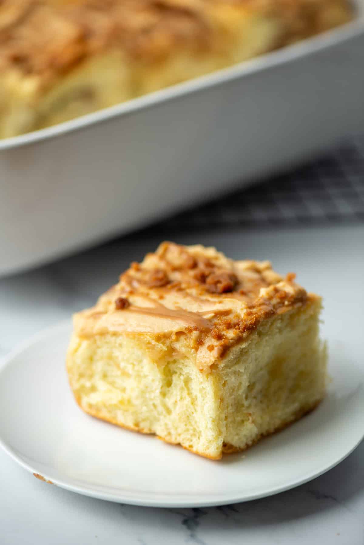 Side view of one Biscoff cinnamon roll on a plate in front of a pan of cinnamon rolls