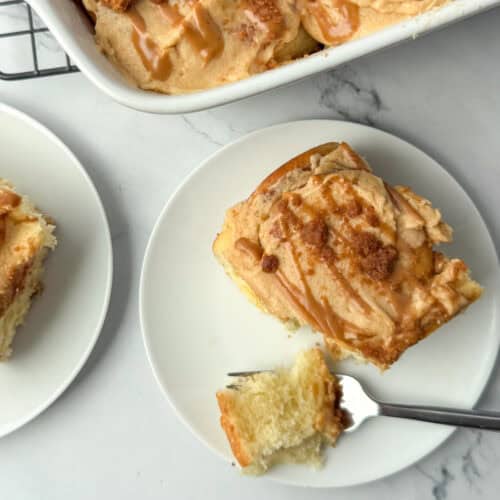 Overhead photo of Biscoff cinnamon roll on a plate with a bite taken out in front of a partial view of a pan of Biscoff cinnamon rolls