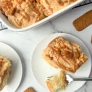 Overhead photo of Biscoff cinnamon roll on a plate with a bite taken out in front of a partial view of a pan of Biscoff cinnamon rolls with a few Biscoff cookies scattered around