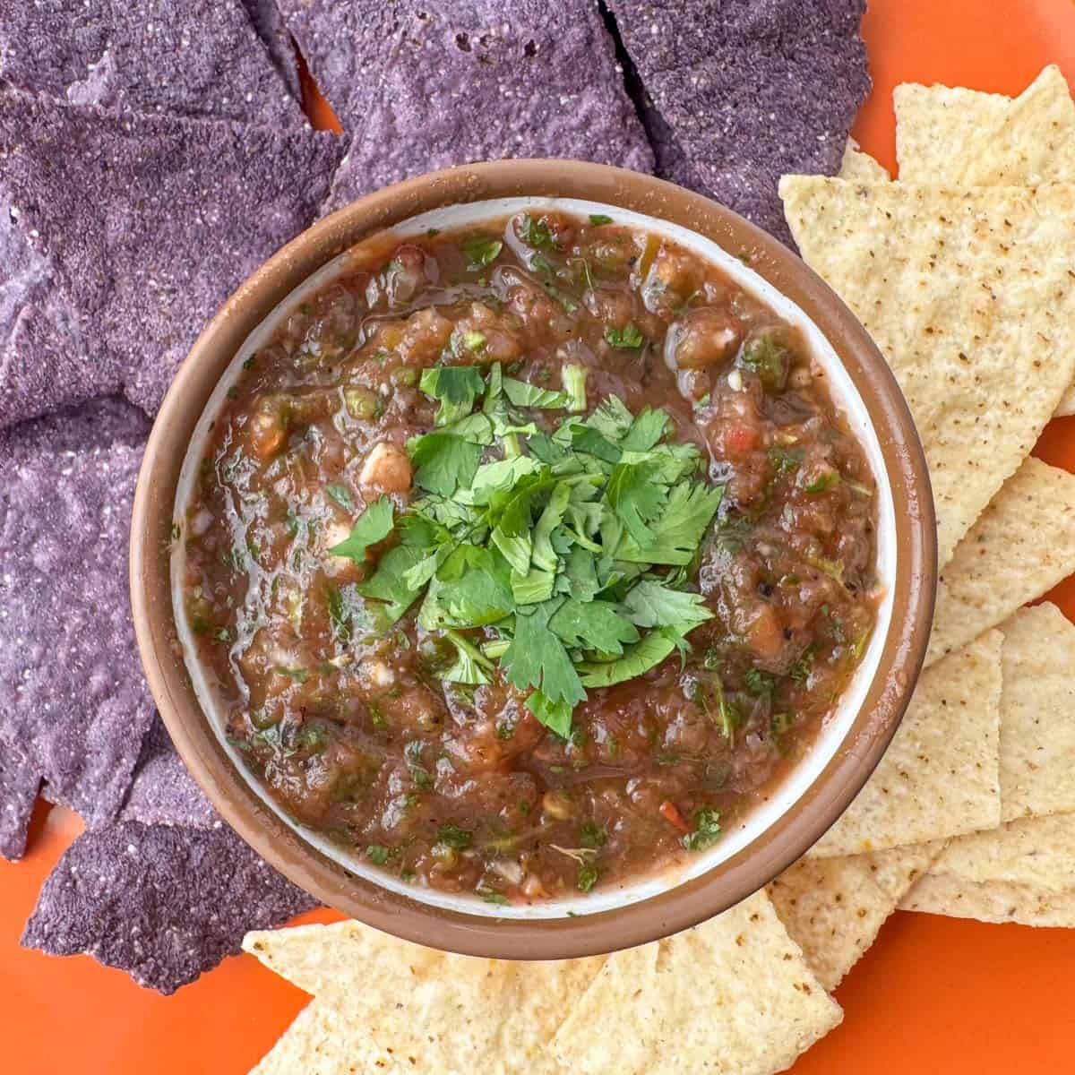 Overhead view of bowl of restaurant style blender salsa made with fire roasted tomatoes on an orange plate with half regular tortilla chips and half blue corn tortilla chips