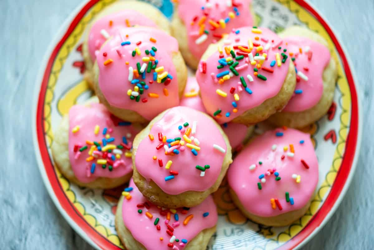 Overhead photo of sour cream cookies with pink frosting and multicolored sprinkles stacked up on a multi colored plate