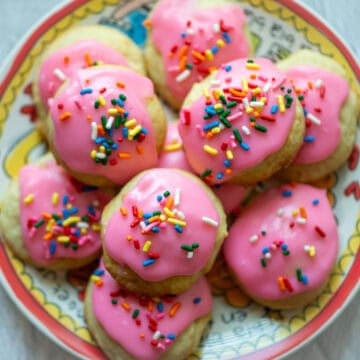 Overhead photo of sour cream cookies with pink frosting and multicolored sprinkles stacked up on a multi colored plate