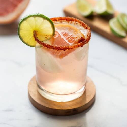 Side view of Paloma mocktail with a Tajin rim garnished with lime and a grapefruit slice with lime slices and a grapefruit in the background