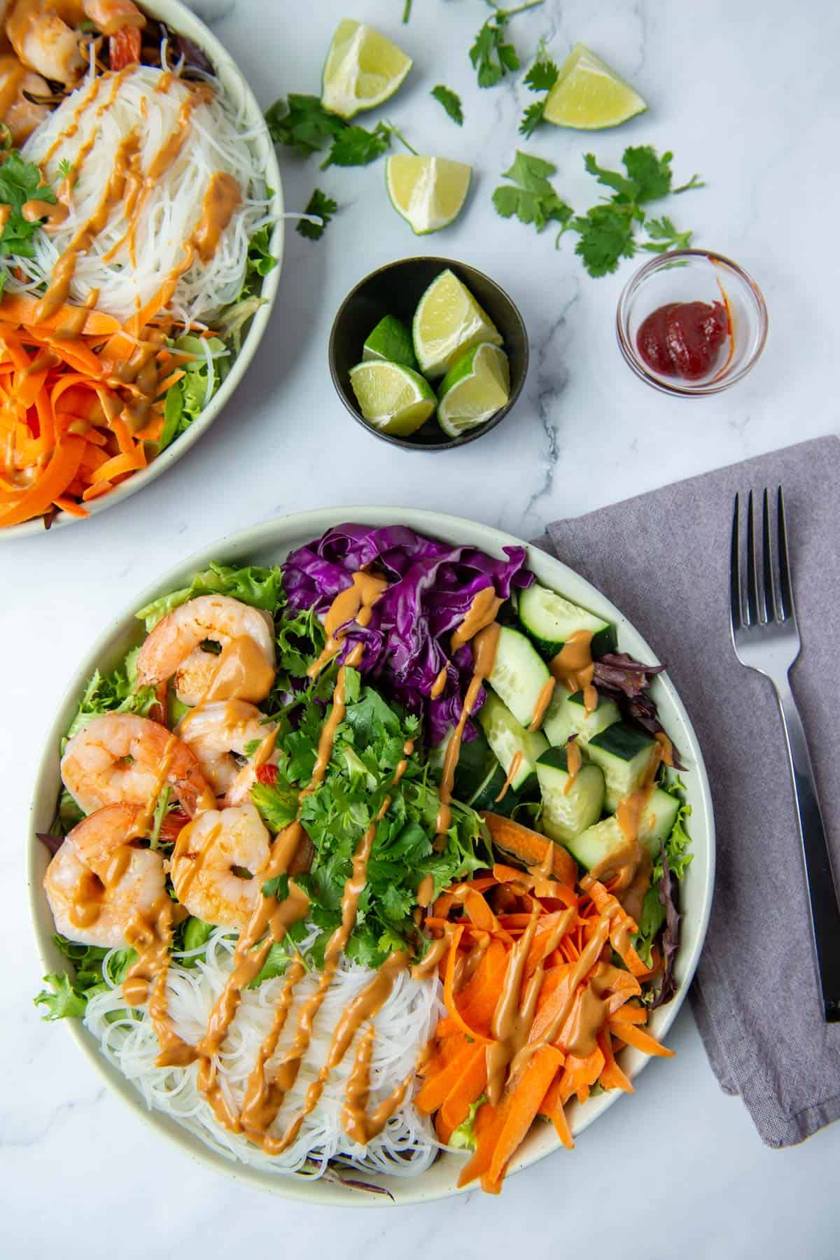 Overhead view of entree size plate of Spring Roll Salad drizzled with peanut dressing, next to a fork and a gray napkin, a bowl of limes, random limes and cilantro, a very small bowl of Gochujang and another plate of Spring Roll Salad