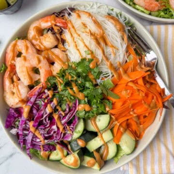 Overhead view of entree size plate of Spring Roll Salad drizzled with peanut dressing, next to a fork and napkin, limes and another plate of Spring Roll Salad
