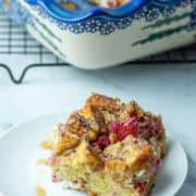 Overhead photo of slice of baked raspberry brioche French toast casserole on a plate with the rest of the baking pan of French toast casserole in the background