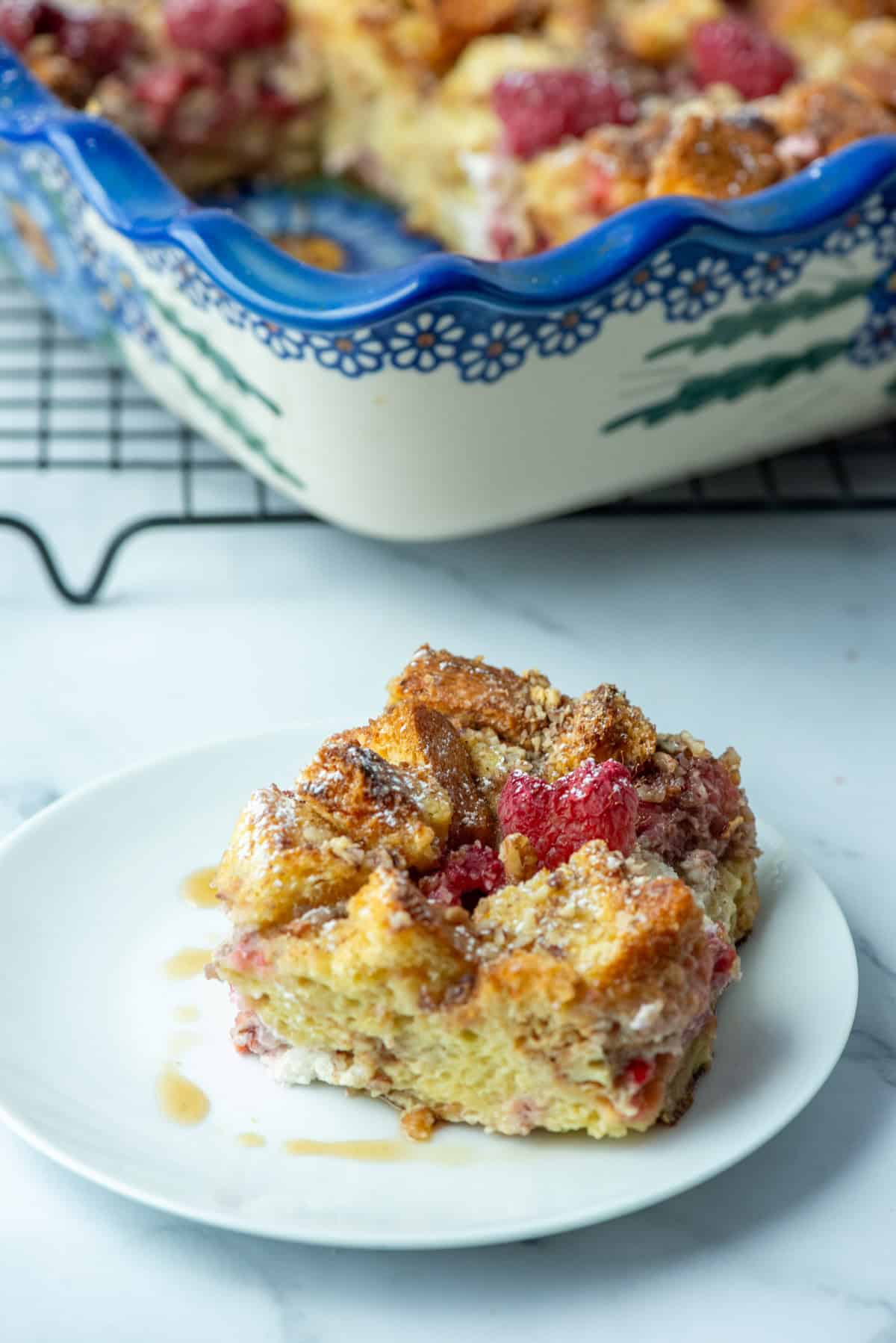 Overhead photo of slice of baked raspberry brioche French toast casserole on a plate with the rest of the baking pan of French toast casserole in the background