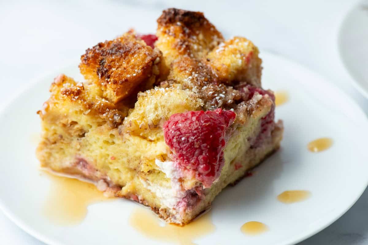 Close up of a slice of Raspberry Brioche French Toast Casserole on a plate with drips of maple syrup. It is cut so that you can see the different layers in the casserole.