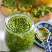 45 degree angle photo of basil pesto in a small, glass jar surrounded by loose basil leaves, a spoon on top of a spoon rest, and small slices of baguette topped with basil pesto and a lemon.