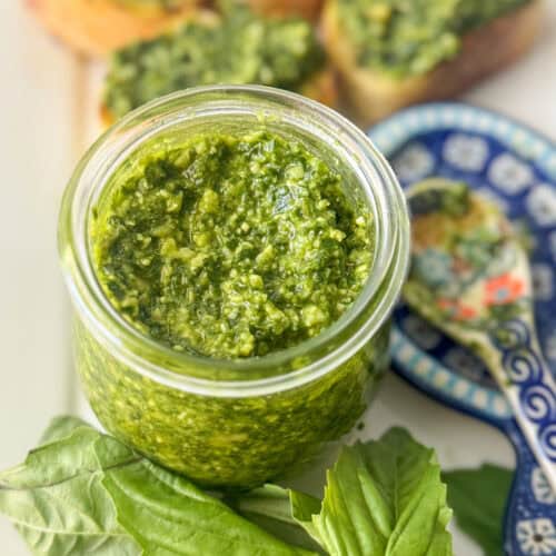 Close up overhead photo of basil pesto in a small, glass jar surrounded by loose basil leaves, a spoon on top of a spoon rest, and small slices of baguette topped with basil pesto.