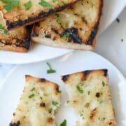 A ripped piece of grilled garlic bread made with a baguette on a white plate in front of a serving plate with the rest of the grilled garlic bread