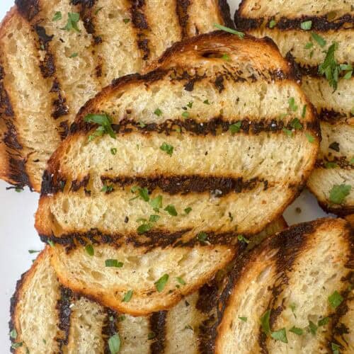 Overhead close up photo of a stack of grilled garlic bread with nice grill char marks and garnished with extra salt and fresh parsley on a serving plate